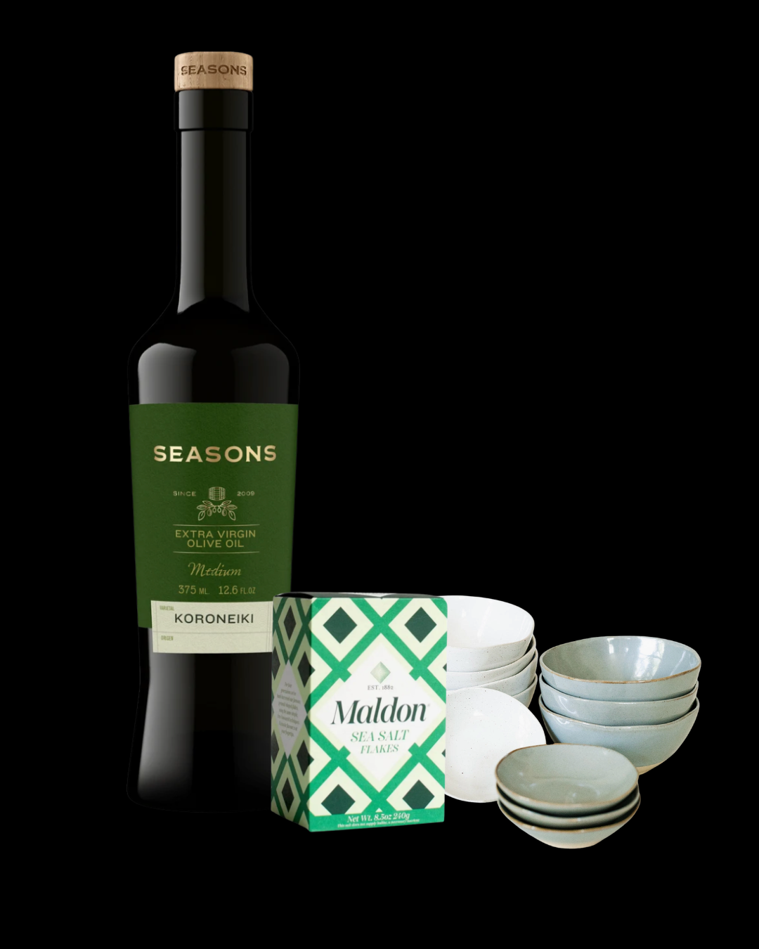 A bottle of Seasons Koroneiki olive oil, a box of Maldon sea salt flakes, and stacks of hand-glazed dipping bowls in gray and ivory